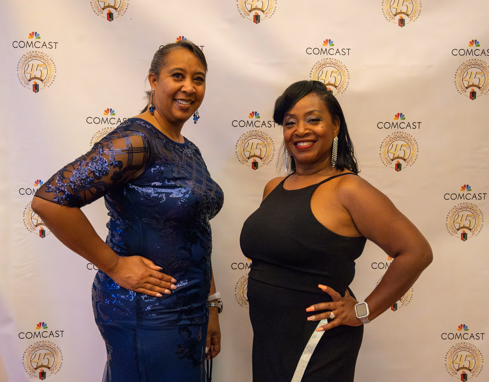 Two guests in blue lace and black gowns pose with hands on hips before the gala logo backdrop
