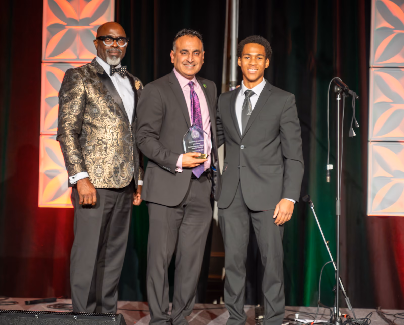 Three guests in suits pose onstage, with the center guest holding a clear award