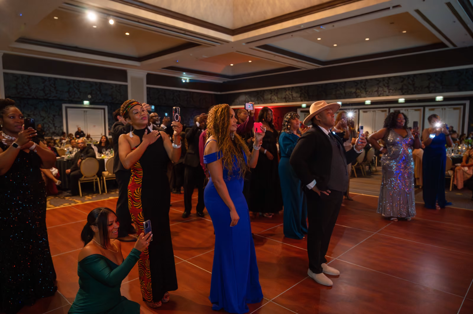 Formally dressed guests gather on the ballroom dance floor, several recording with their phones