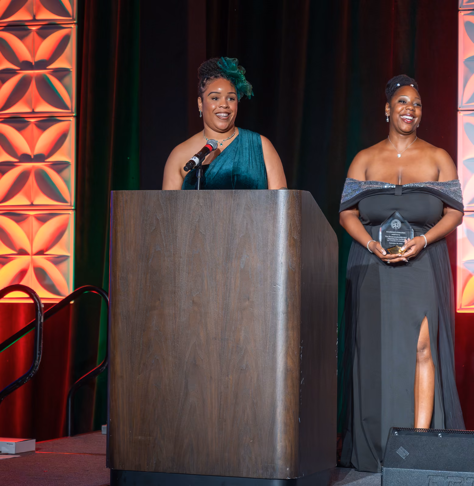 A speaker in a green gown stands at a wooden podium beside a smiling guest holding an award