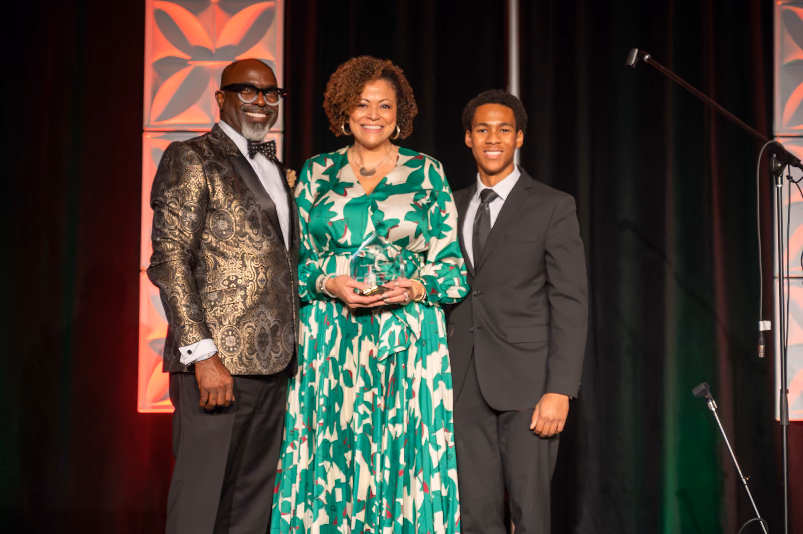 A guest in a green and cream patterned dress holds an award between two suited guests onstage