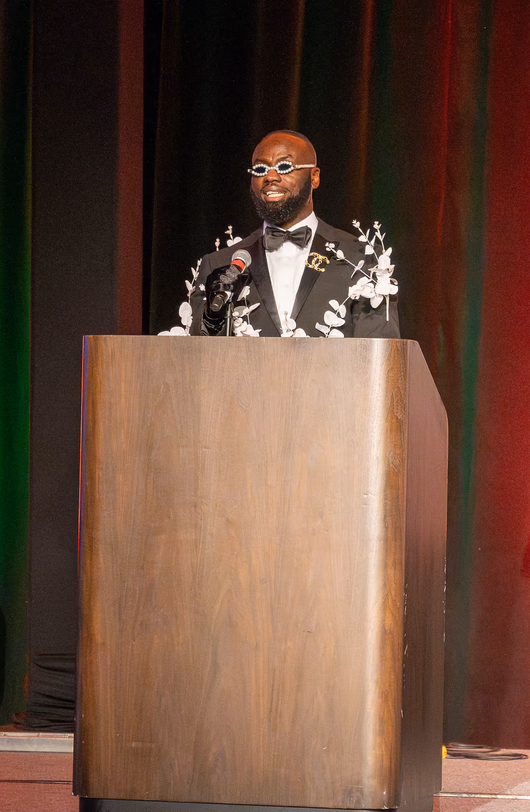 A speaker wearing decorative glasses and a tuxedo with white floral accents stands at a wooden podium