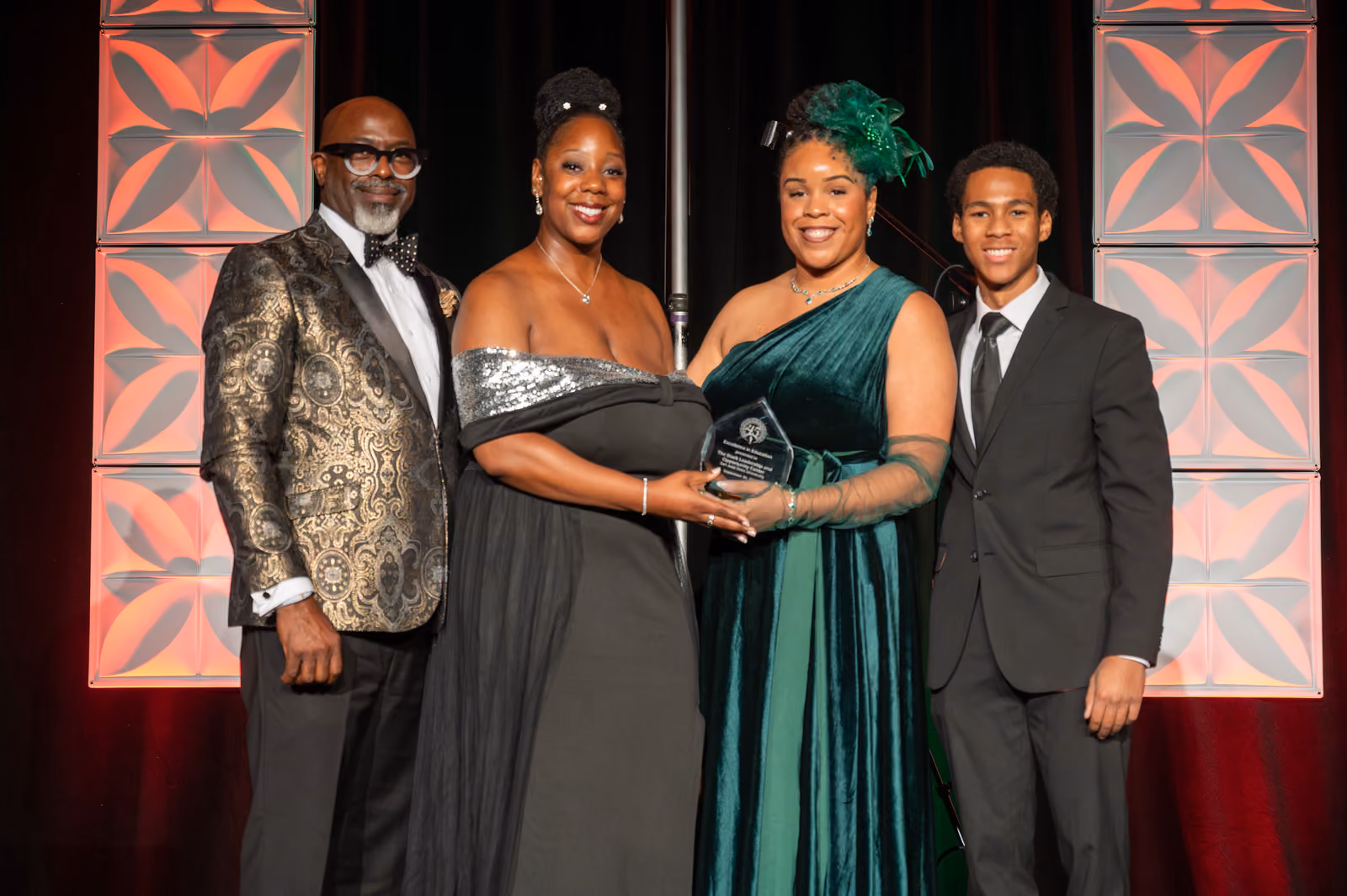 Four formally dressed guests pose onstage, with two guests at the center holding a clear award