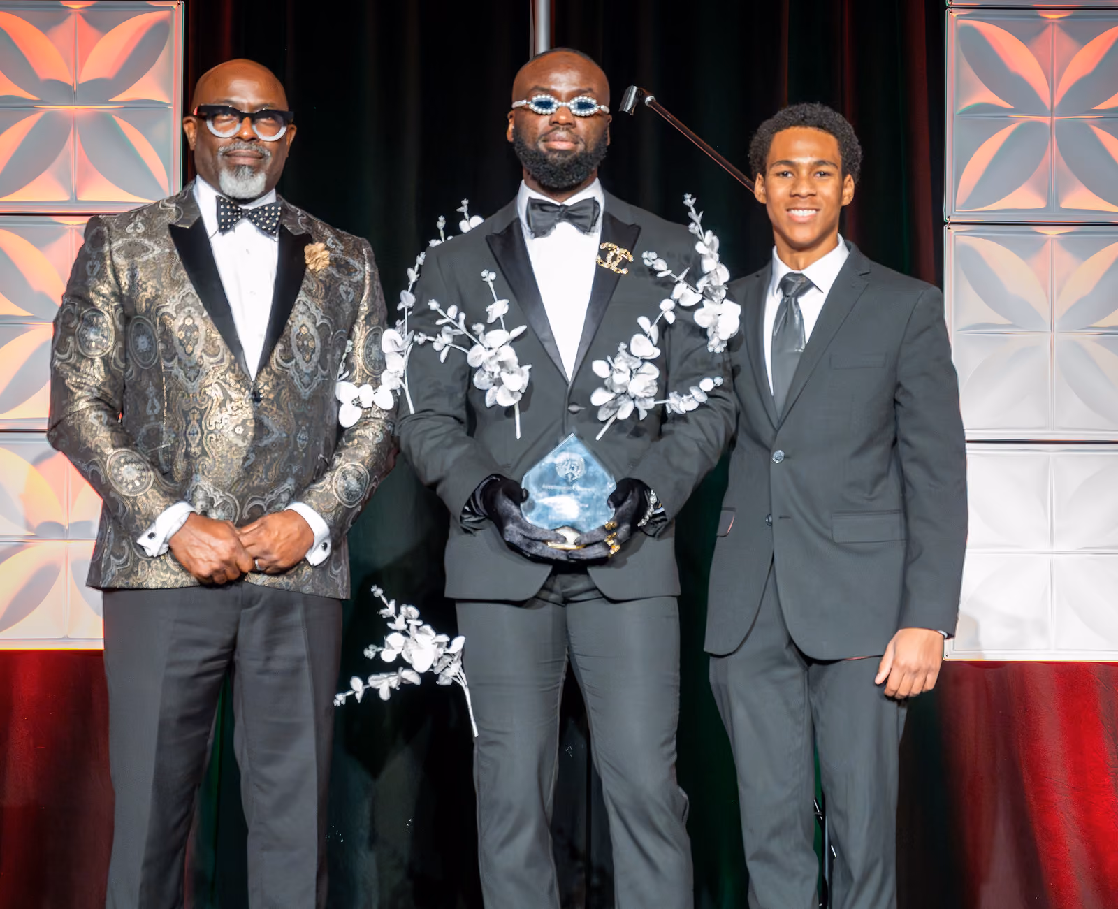 Three guests pose onstage, with an award held by the center guest in a tuxedo with white floral accents