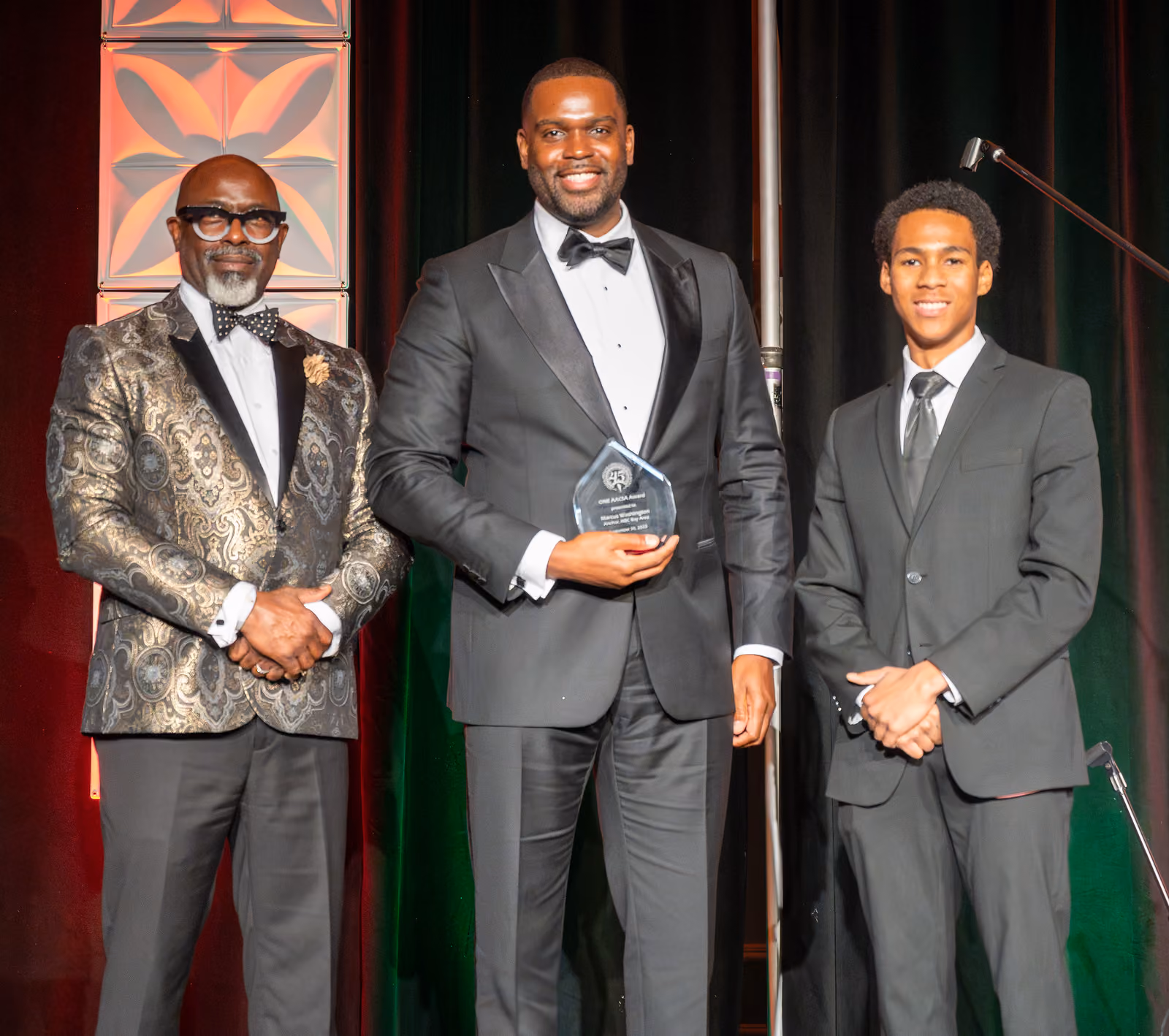 Three guests in formal suits pose before black stage curtains, with the center guest holding an award