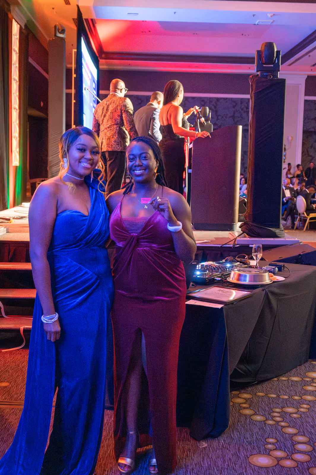 Two guests in blue and burgundy gowns pose beside an audio equipment table near the ballroom stage