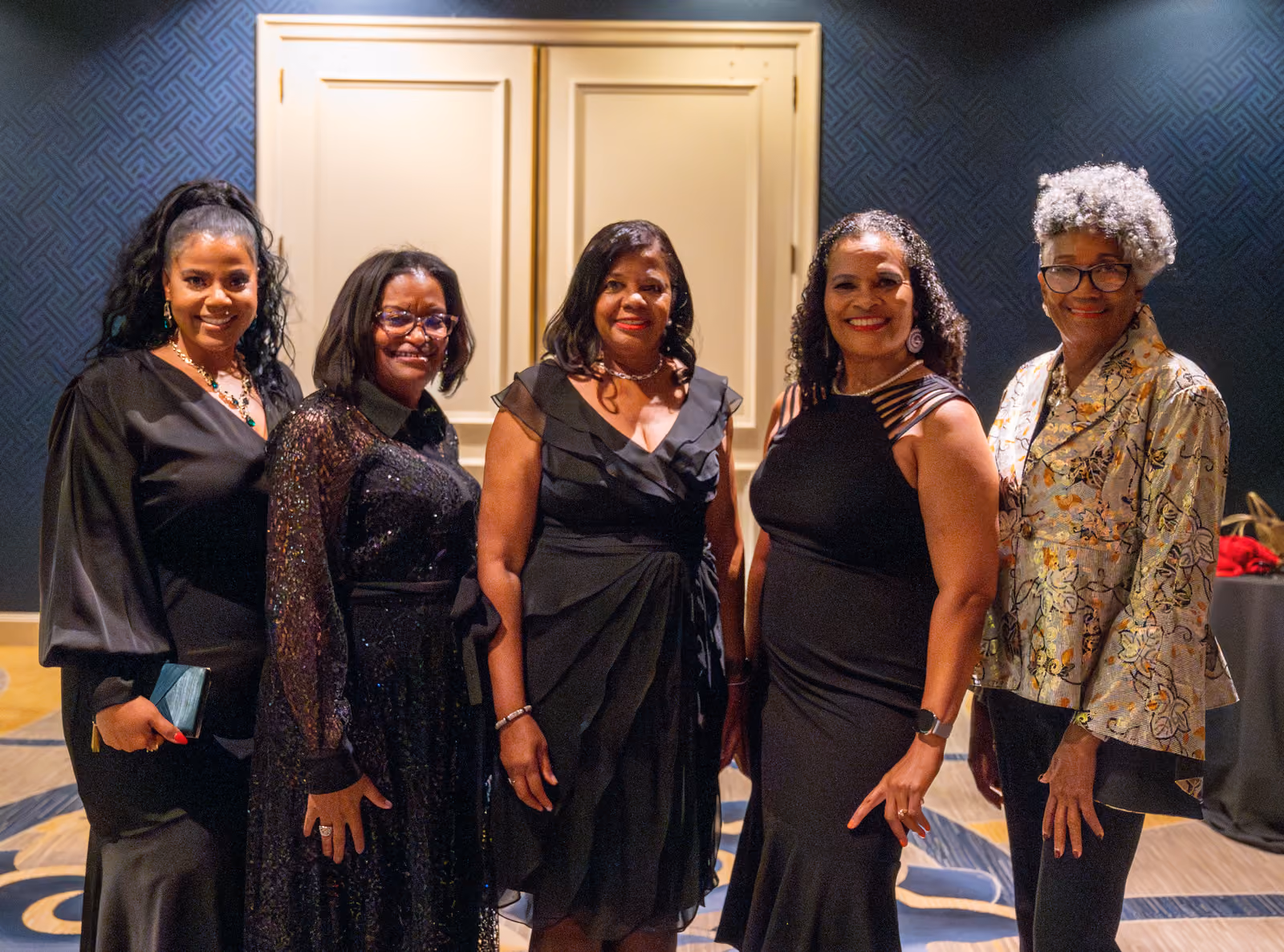 Five smiling guests in formal attire pose together in a hallway before cream double doors