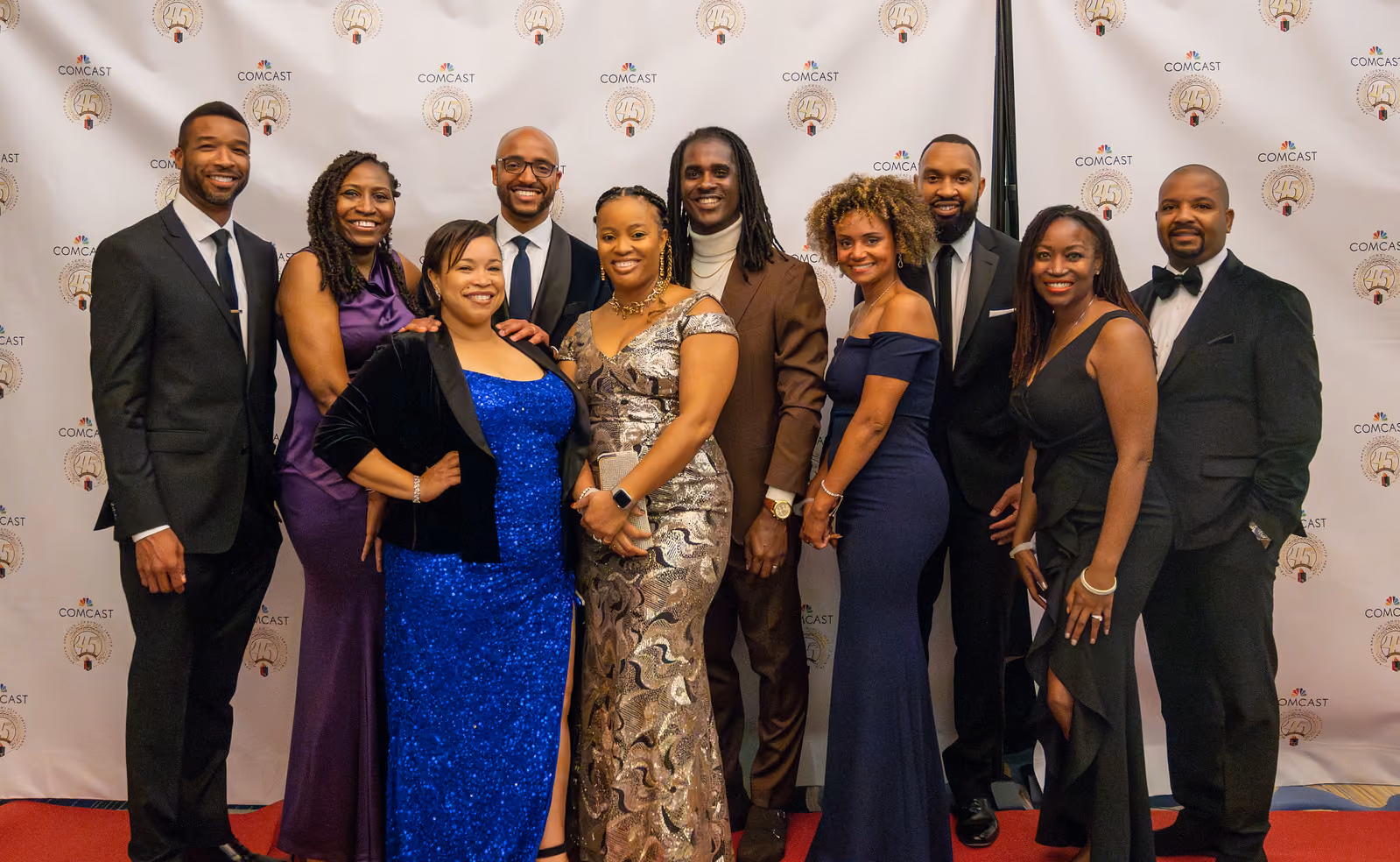 Ten guests in formal gowns and suits pose together on a red carpet before the gala logo backdrop