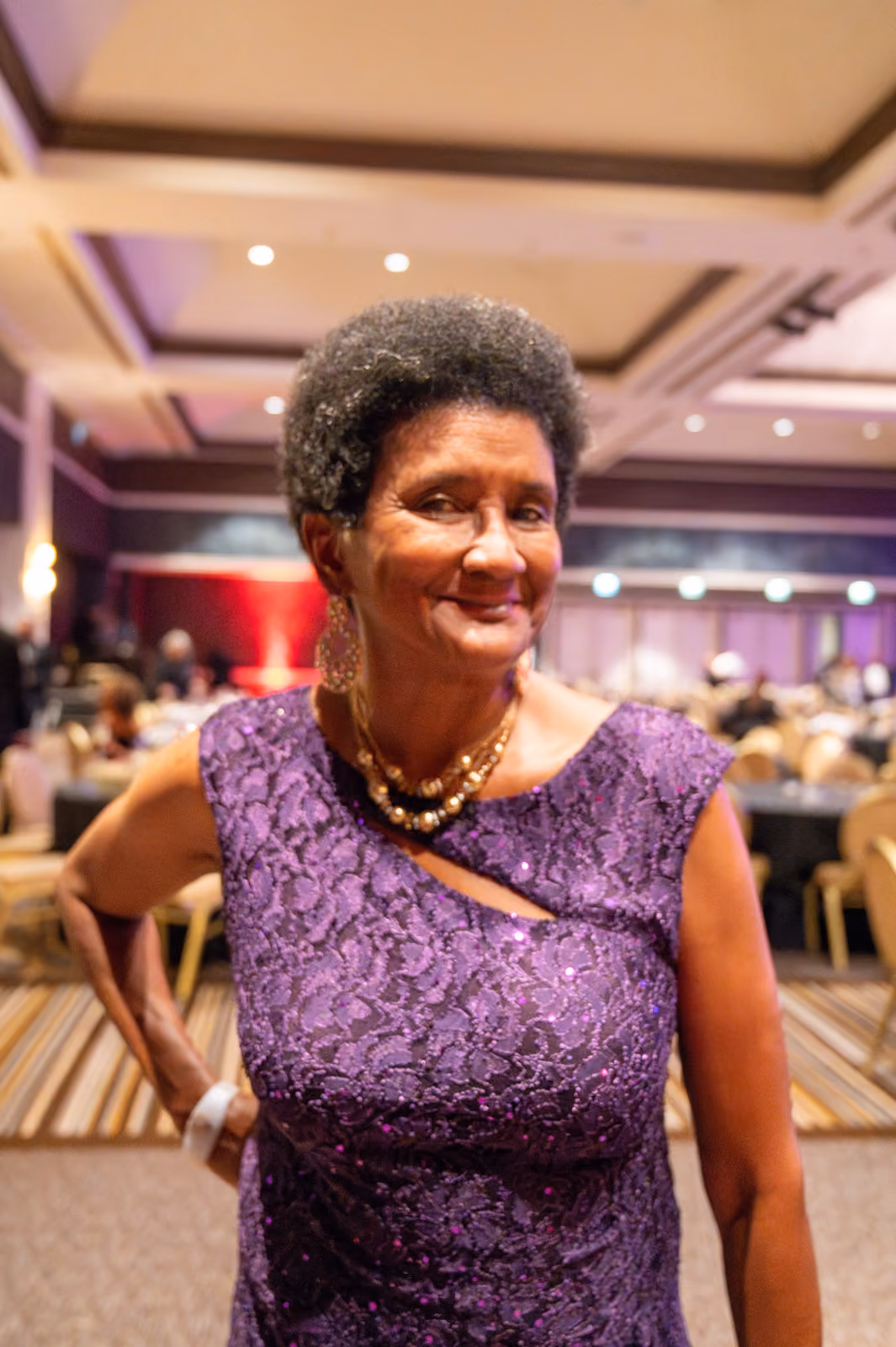 A smiling guest in a purple lace dress and gold jewelry poses in the ballroom near banquet tables