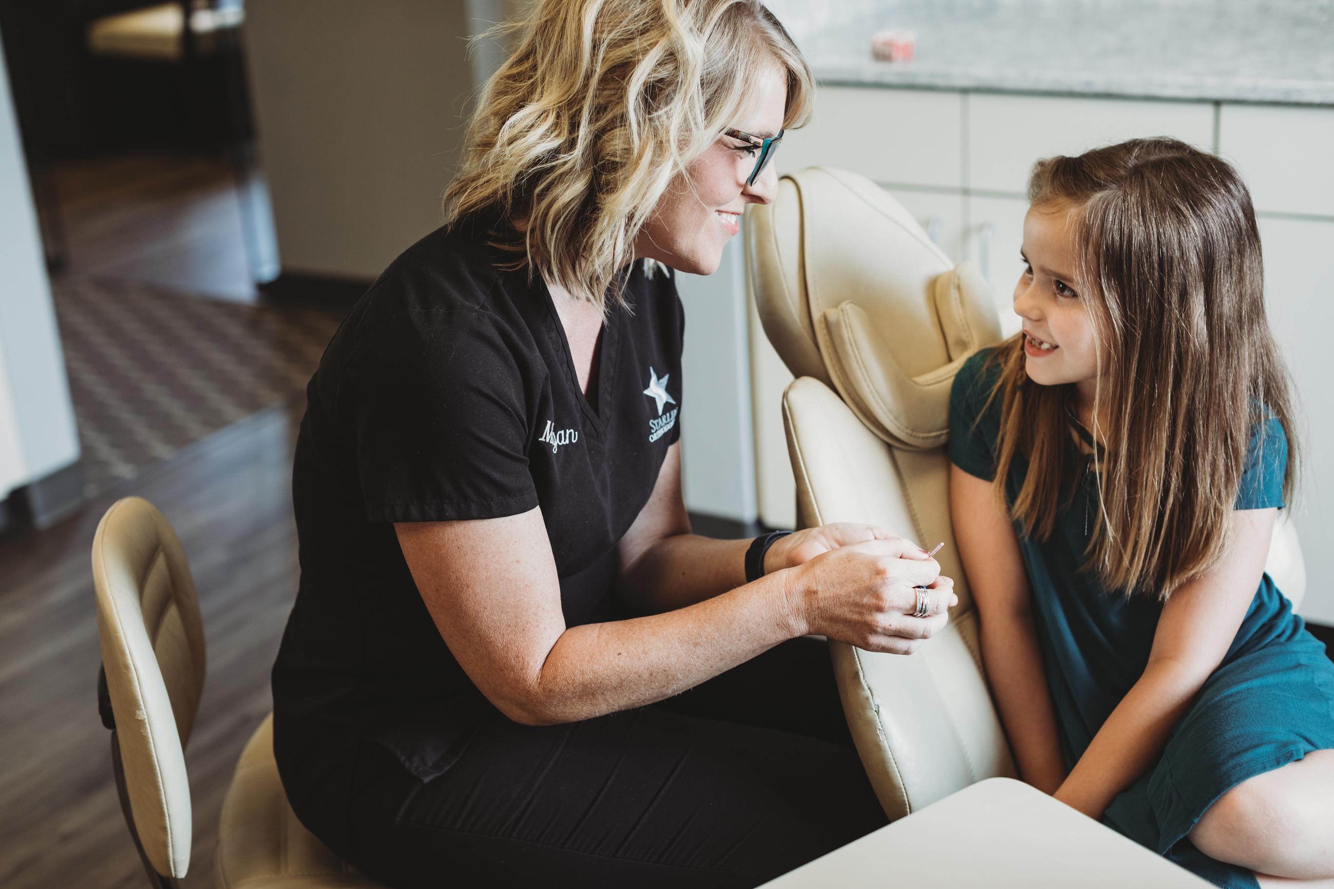 A child is smiling brightly in a welcoming orthodontic office, with a friendly clinician nearby, showcasing the importance of pediatric dentistry and oral health. This positive experience highlights the dedication to providing expert care for children's teeth in a warm environment.
