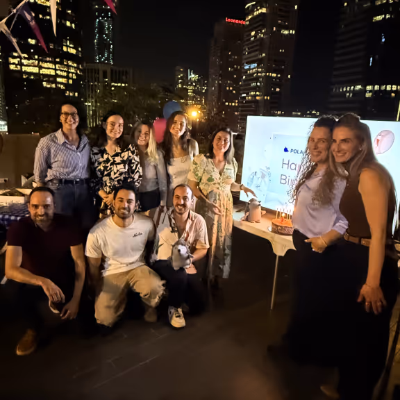 Group of ten people posing at a rooftop birthday party at night with city buildings lit in the background and birthday cake with candles on a table.