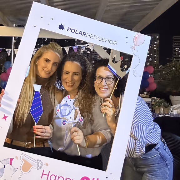 Three women smiling and posing behind a Polar Hedgehog branded photo frame with party props including a crown, tie, and scooter.