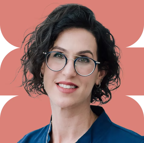 Smiling woman with short curly dark hair, wearing round glasses and a dark blue top against a coral background.