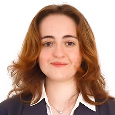Portrait of a woman with shoulder-length auburn hair wearing a dark blazer and white collared shirt against a peach background.