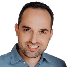 Smiling man with short dark hair, stubble, and a blue collared shirt against a pale yellow background.