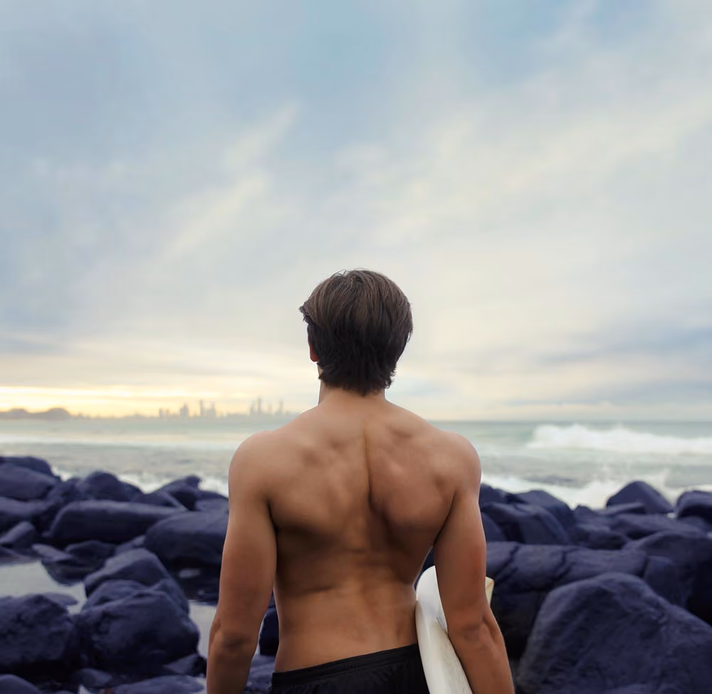Shirtless man holding a surfboard looks at the ocean from rocky shore during cloudy sunset.
