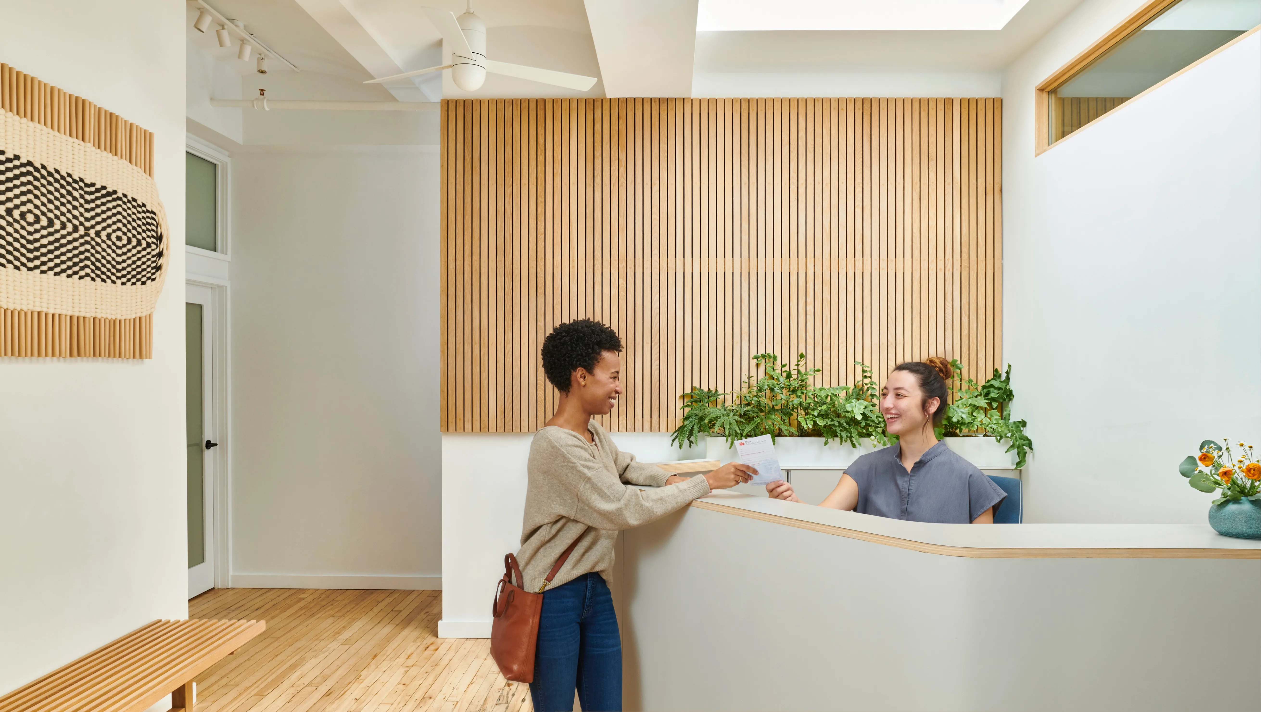 A patient conferring with a staff member in a skylit reception area, with light wood slats and plants in the background, at Olo Acupuncture in New York City.