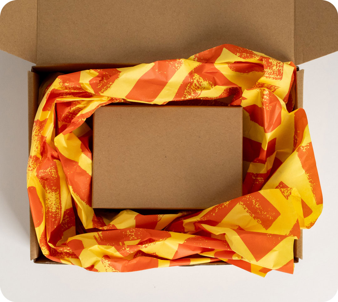 Open cardboard box with a smaller box inside, surrounded by crumpled orange and yellow wrapping paper.