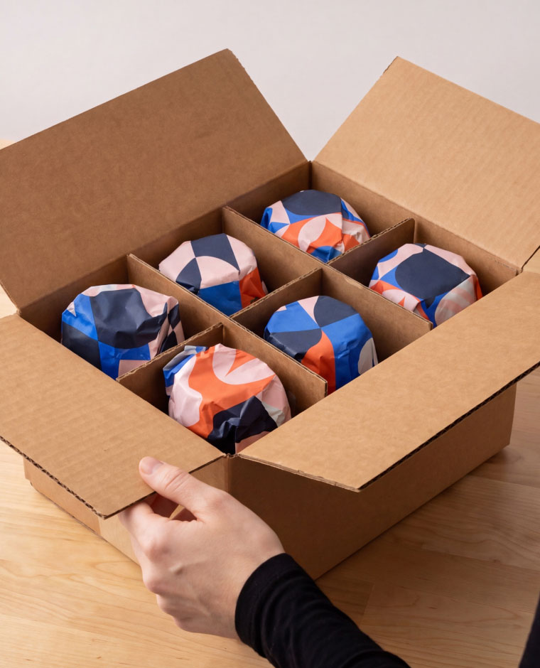 Hand holding open a cardboard box with six compartments, each holding a wrapped item with colorful patterns.