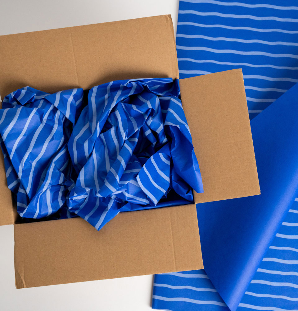 Open cardboard box filled with crumpled blue and white striped packing paper, next to sheets of similar paper.