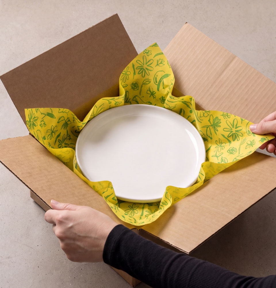 Person unpacking a white ceramic plate wrapped in yellow floral-patterned packing paper slip sheets inside a cardboard box.