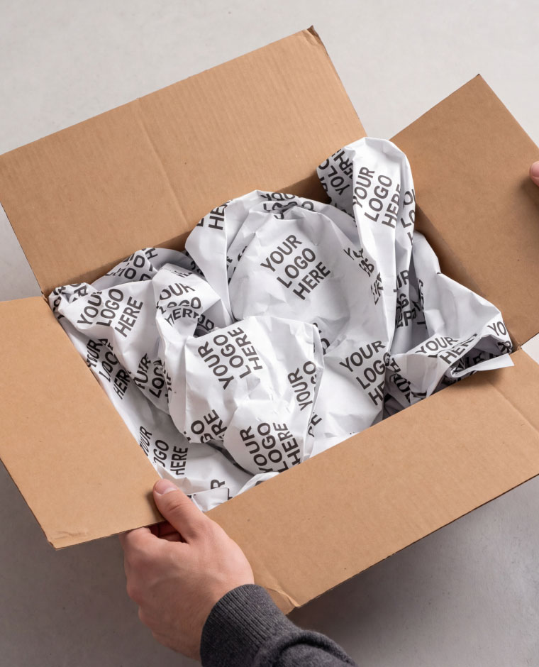 Hands holding open a cardboard box filled with crumpled white packing paper printed with "Your Logo Here."