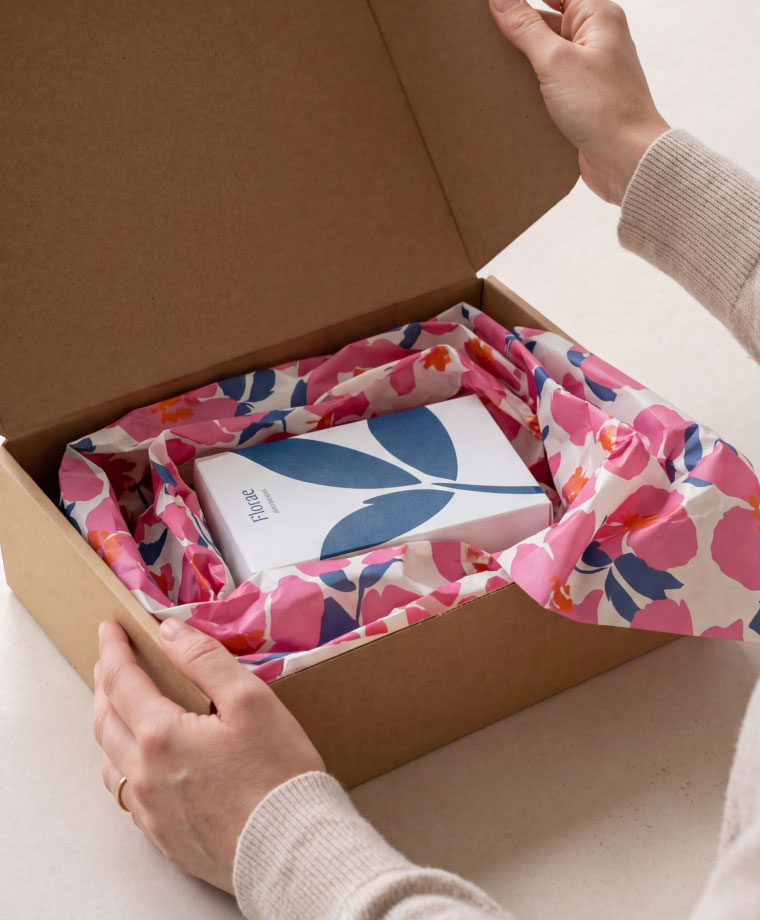 Hands opening a cardboard box with floral satinpack paper and a white box with blue leaf design inside.