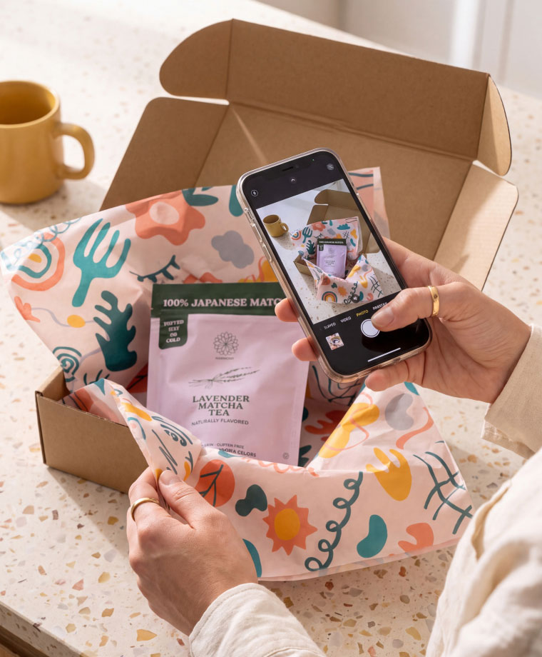 Hands holding colorful tissue around boxed lavender matcha tea while taking photo with smartphone.