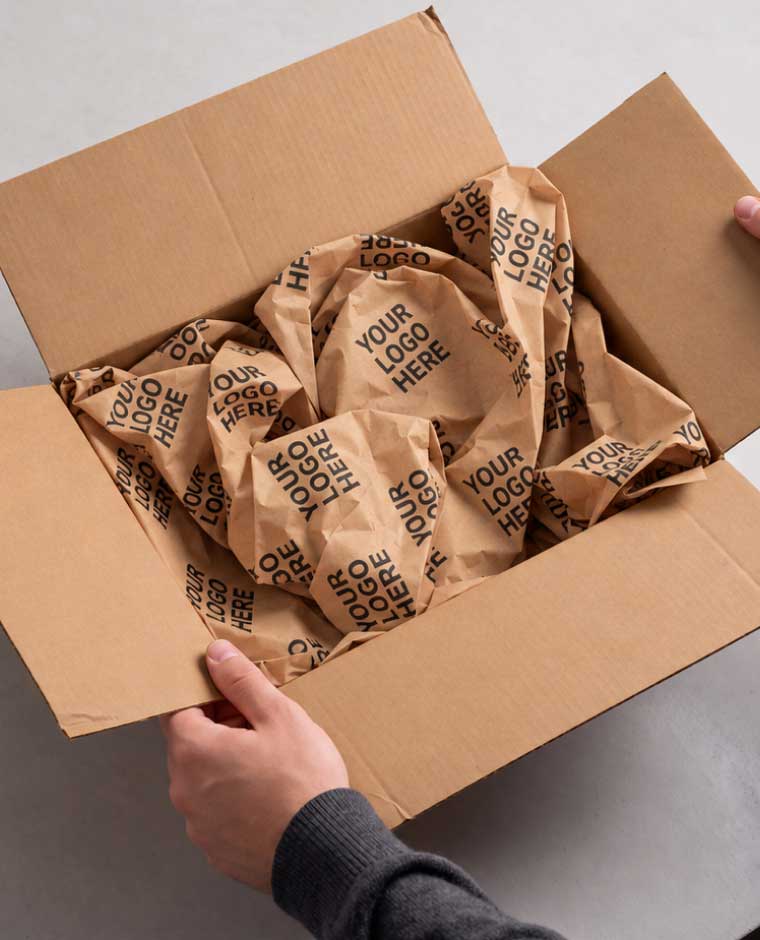 Hands holding open a cardboard box filled with crumpled white packing paper printed with "Your Logo Here."