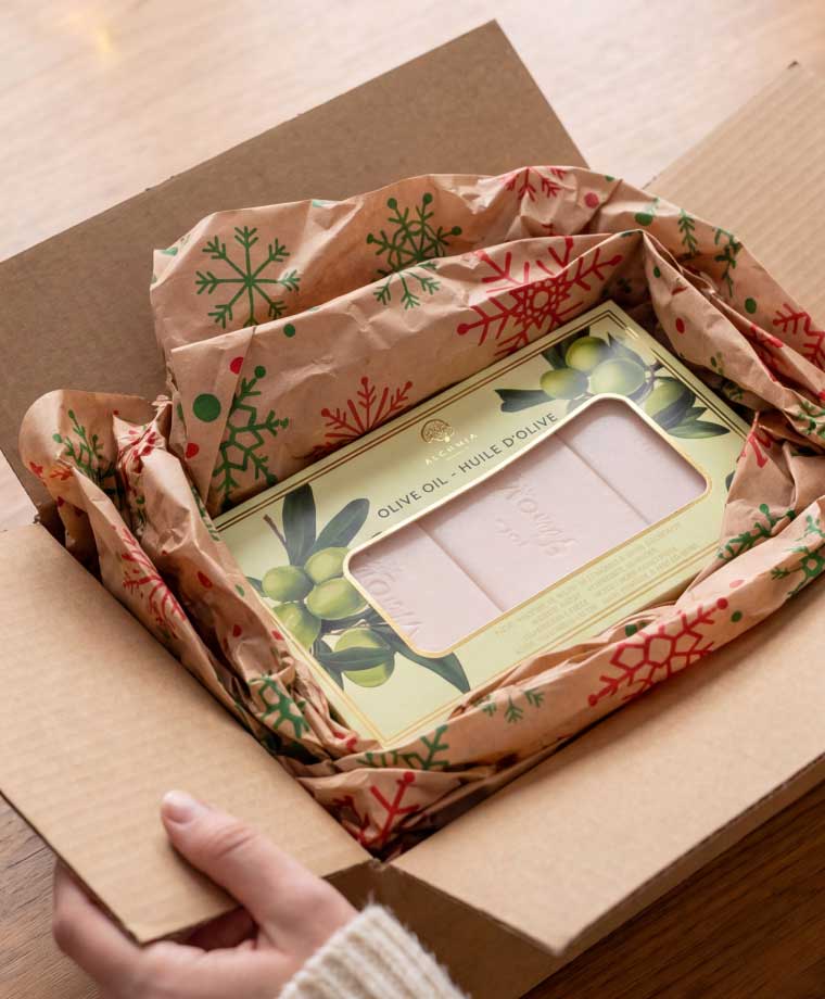 A hand opening a cardboard box containing a gift-wrapped package with red and green snowflake patterns, revealing a box of olive oil soap bars inside a windowed box.