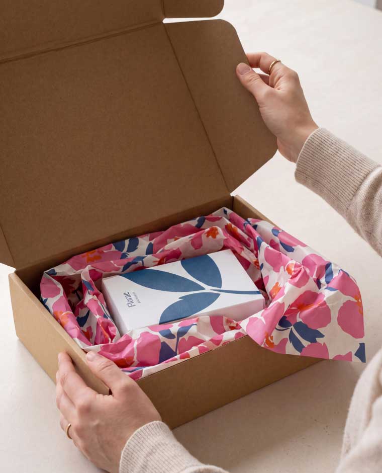 A person opening a brown cardboard box lined with pink, blue, and white floral-patterned fabric, revealing a white product box labeled 'Florae' with a large blue leaf design on the top.