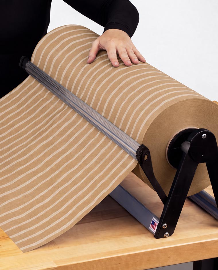 A person holding a large roll of brown kraft paper with white vertical stripes on a tabletop dispenser with a wooden surface.