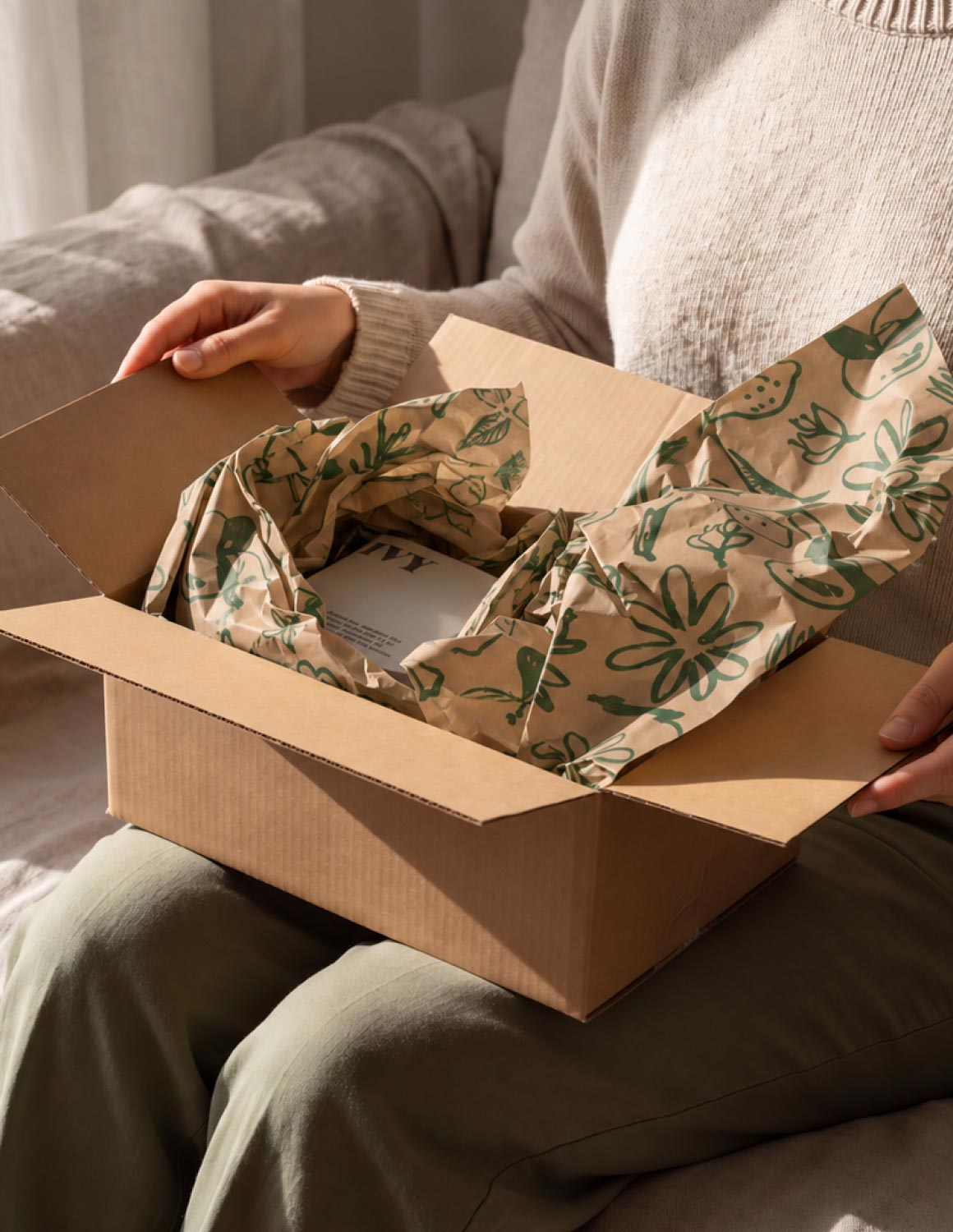 Person sitting with a cardboard box on their lap, opening it to reveal crumpled kraft paper with green plant illustrations used as packing material inside the box.