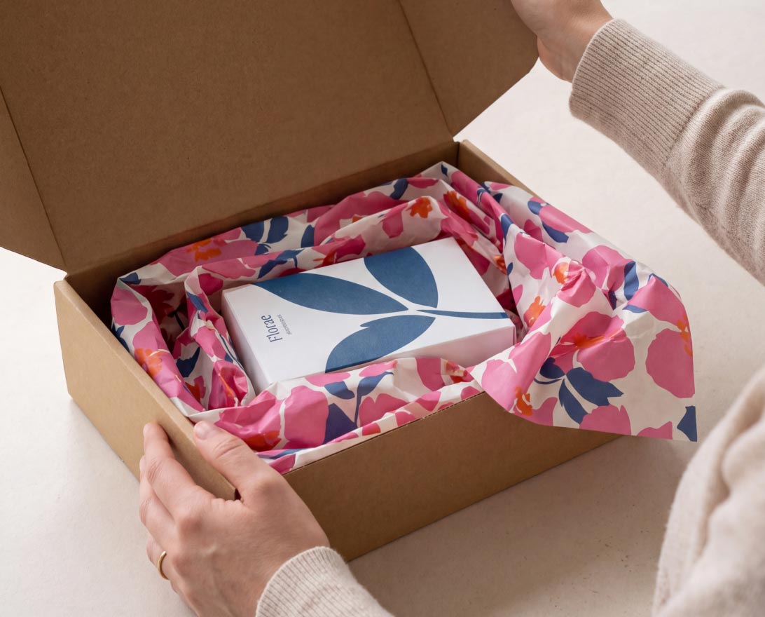 Hands holding open a cardboard box filled with crumpled white packing paper printed with "Your Logo Here."