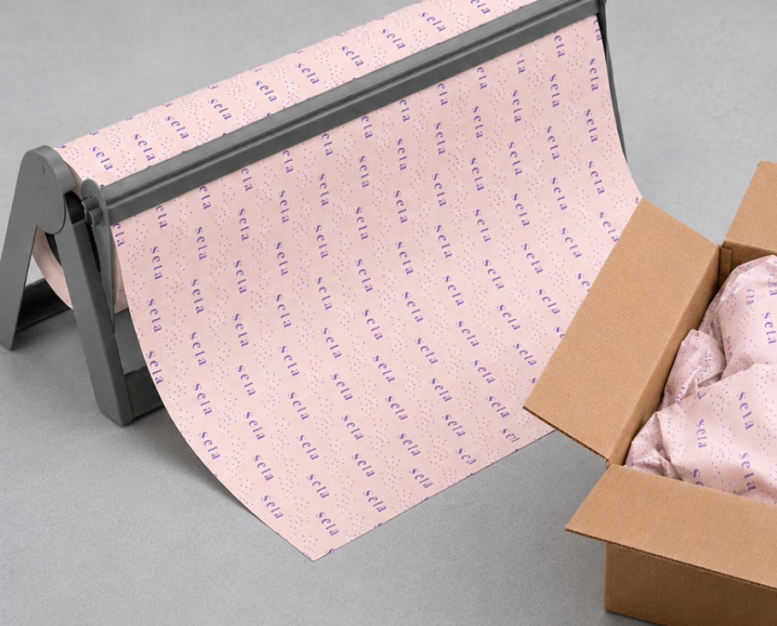 Roll of pink branded wrapping paper with the word "sela" printed in purple repeatedly, partially unrolled from a black stand next to a cardboard box containing crumpled sheets of the same wrapping paper.