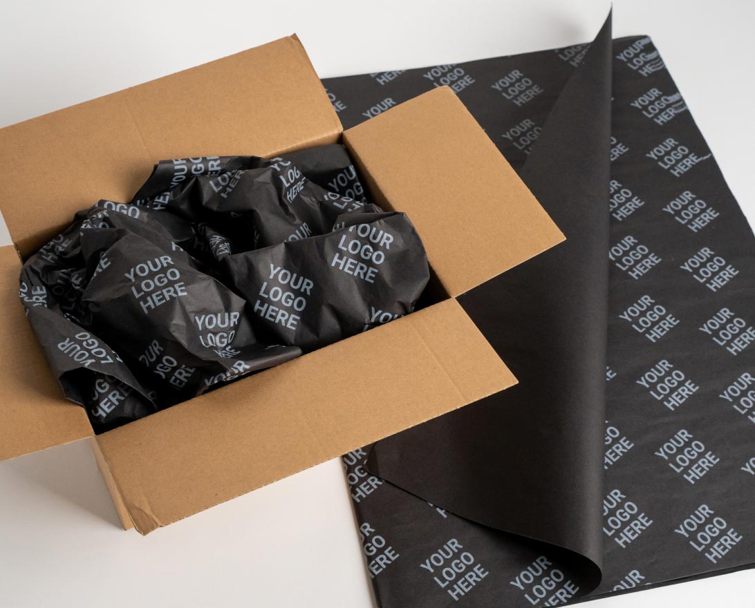 Open brown cardboard box filled with crumpled black packing paper printed repeatedly with the phrase 'YOUR LOGO HERE' in white, next to a flat sheet of the same packing paper partially folded.