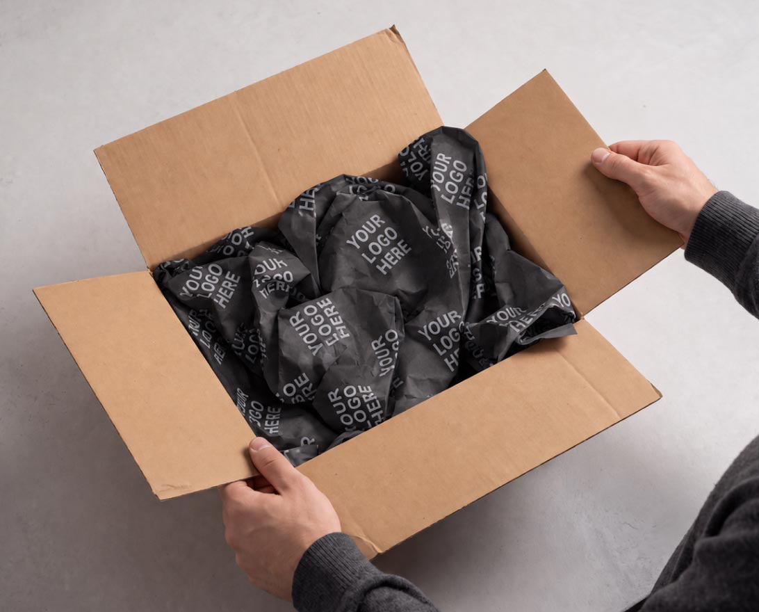 Adult hands holding open a brown cardboard box filled with crumpled black packing paper printed repeatedly with the phrase 'YOUR LOGO HERE'.