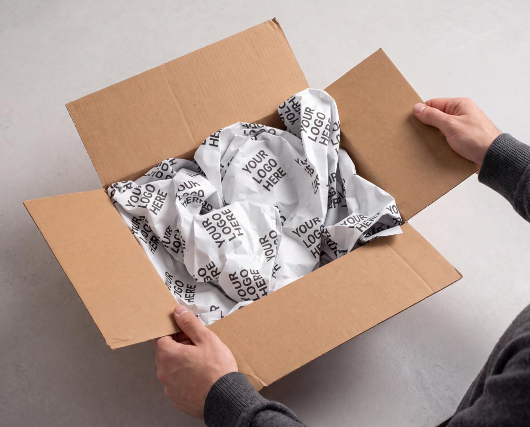 Adult hands holding open a brown cardboard box filled with crumpled white packing paper printed repeatedly with the phrase 'YOUR LOGO HERE'.