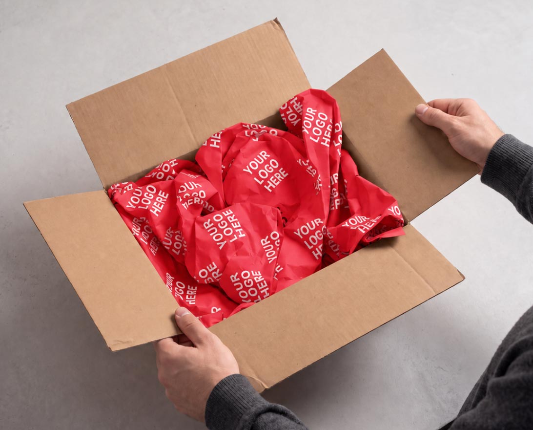 Adult hands holding open a brown cardboard box filled with crumpled red packing paper printed repeatedly with the phrase 'YOUR LOGO HERE'.