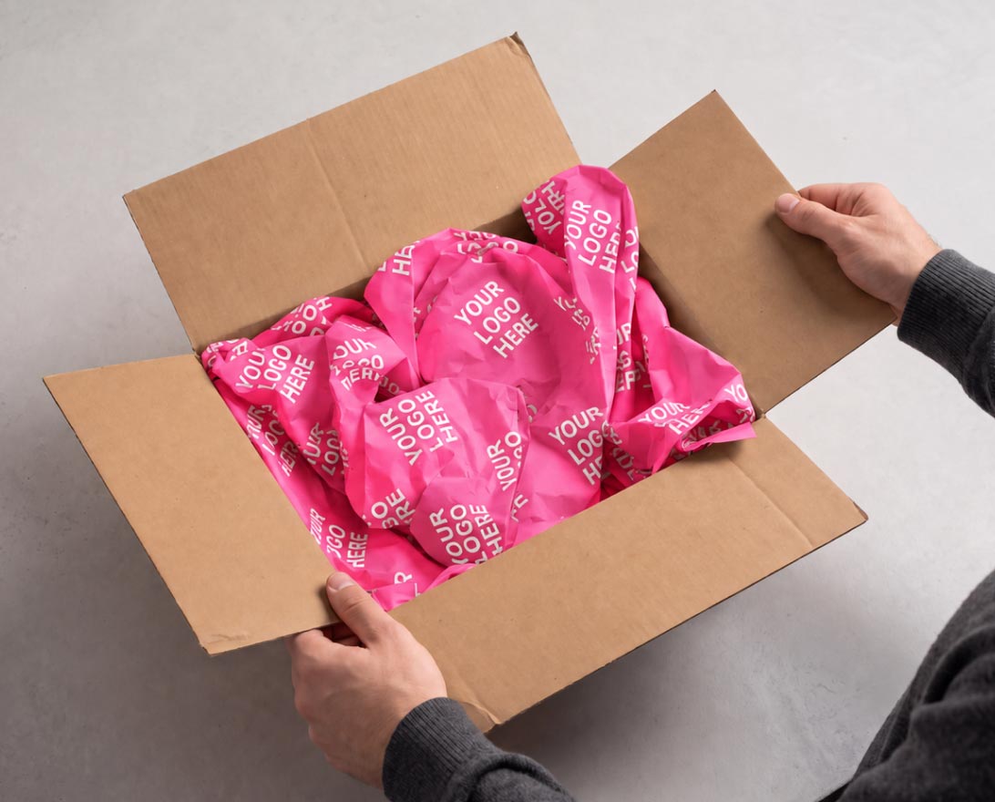 Adult hands holding open a brown cardboard box filled with crumpled hot pink packing paper printed repeatedly with the phrase 'YOUR LOGO HERE'.