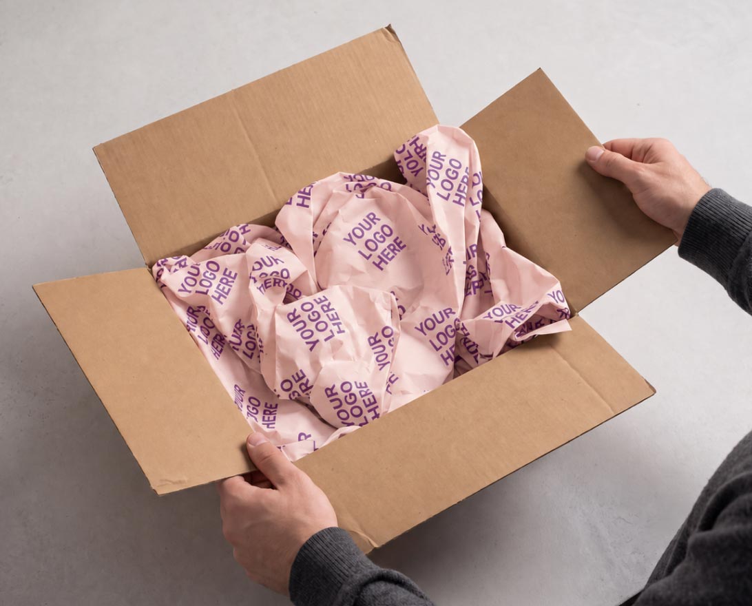 Adult hands holding open a brown cardboard box filled with crumpled light pink packing paper printed repeatedly with the phrase 'YOUR LOGO HERE'.