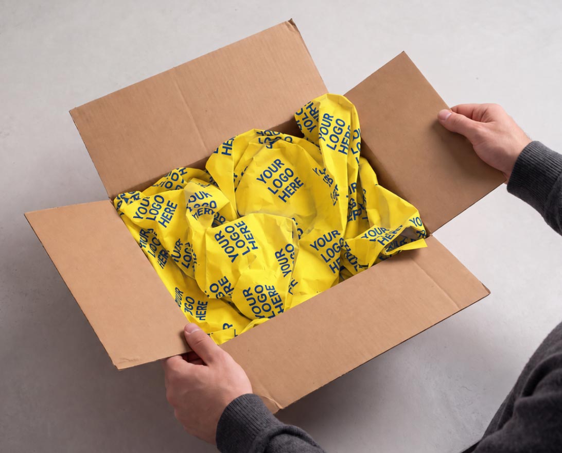 Adult hands holding open a brown cardboard box filled with crumpled yellow packing paper printed repeatedly with the phrase 'YOUR LOGO HERE'.