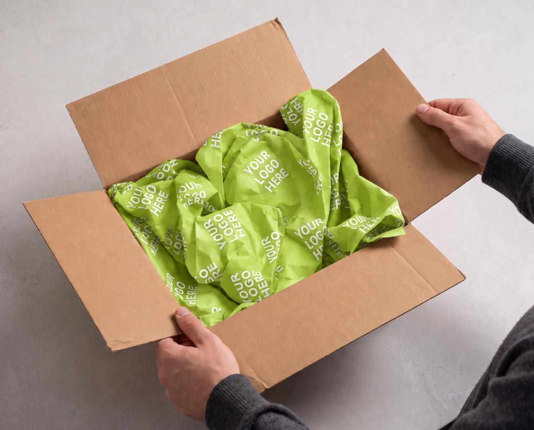 Adult hands holding open a brown cardboard box filled with crumpled green packing paper printed repeatedly with the phrase 'YOUR LOGO HERE'.