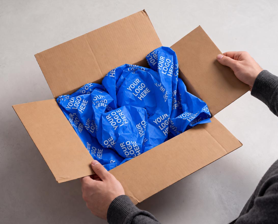 Adult hands holding open a brown cardboard box filled with crumpled blue packing paper printed repeatedly with the phrase 'YOUR LOGO HERE'.