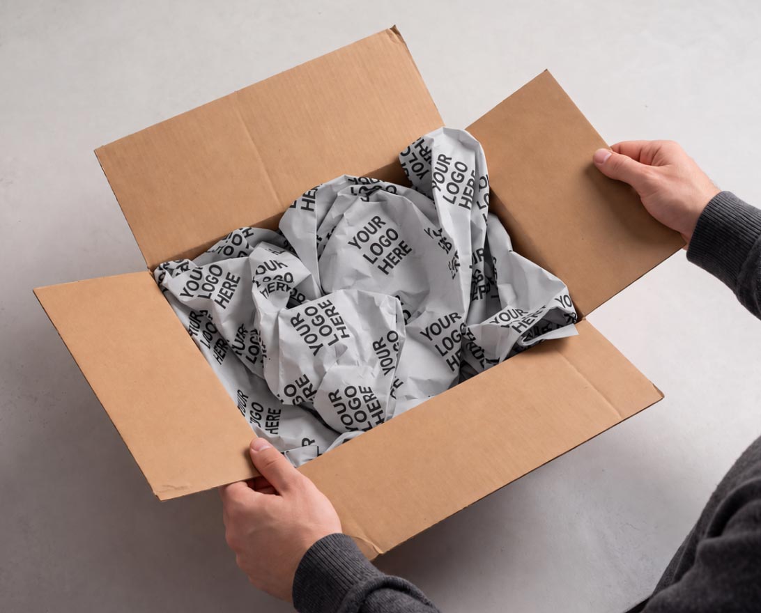 Adult hands holding open a brown cardboard box filled with crumpled gray packing paper printed repeatedly with the phrase 'YOUR LOGO HERE'.