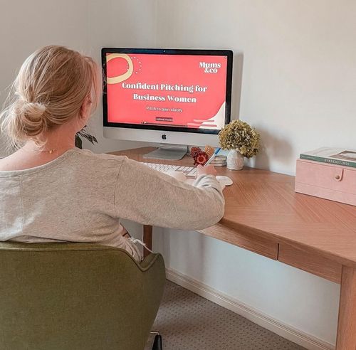 Woman with blonde hair in a bun sitting at a wooden desk, taking notes while watching a computer screen displaying 'Confident Pitching for Business Women' course.