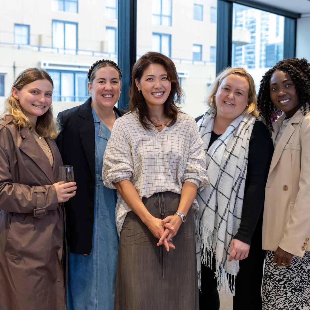 Group of five smiling women standing closely together in a modern indoor space with large windows.