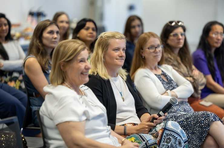 Smiling woman with shoulder-length blonde hair wearing a purple and blue patterned top, sitting in front of a window.