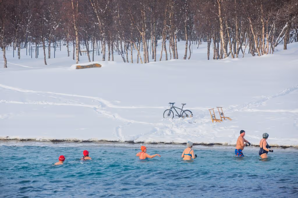 Adventurous winter swimmers taking a dip in an icy lake surrounded by snow, with chairs and a bike visible on the snowy shore.