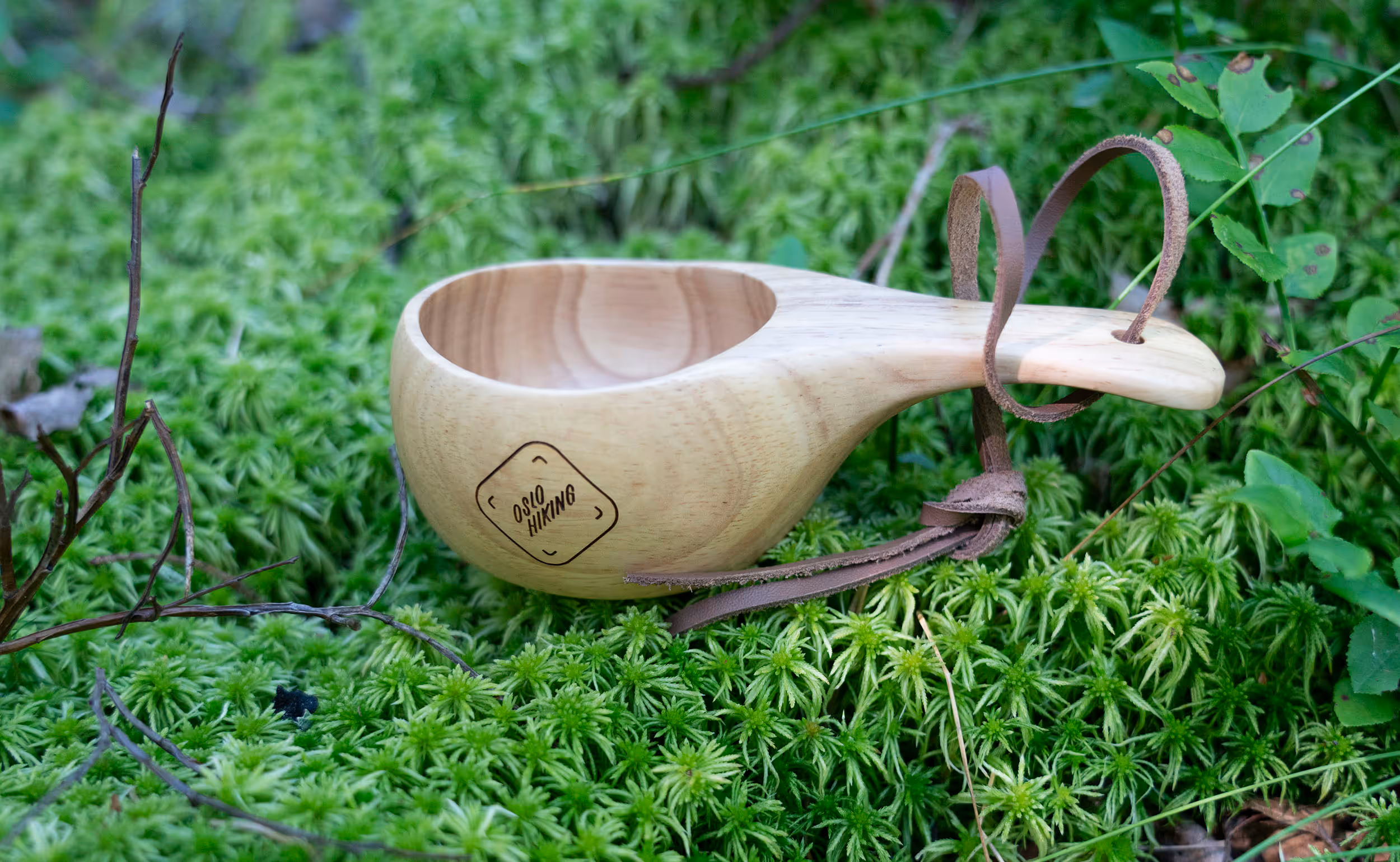 A handcrafted wooden cup engraved with the "Oslo Hiking" logo, resting on a bed of vibrant green moss. A leather strap is attached to the handle, showcasing a practical and rustic design.